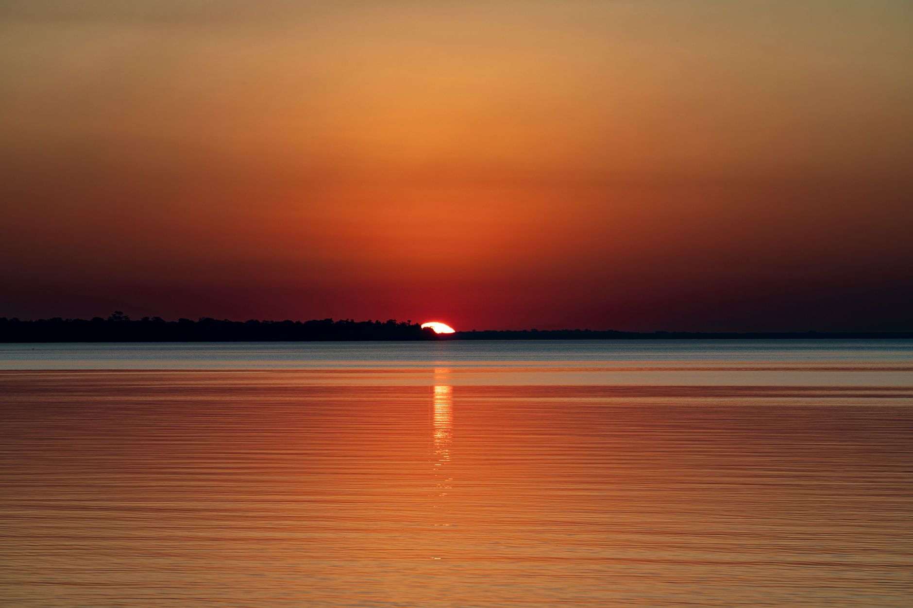 A calm lake at sunset with forested mountains visible in the background along the shoreline