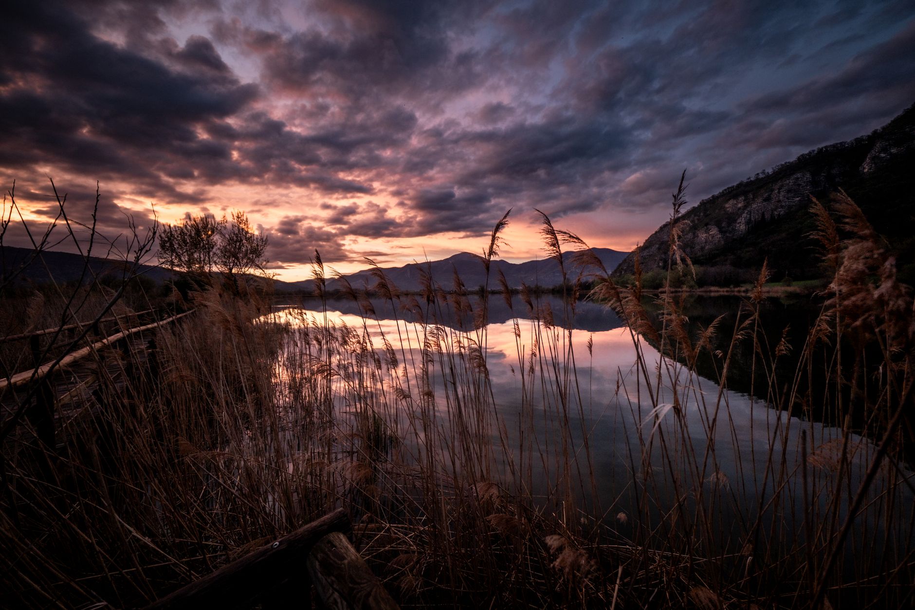 Calm lake shoreline at sunset with mountain silhouettes visible across the water